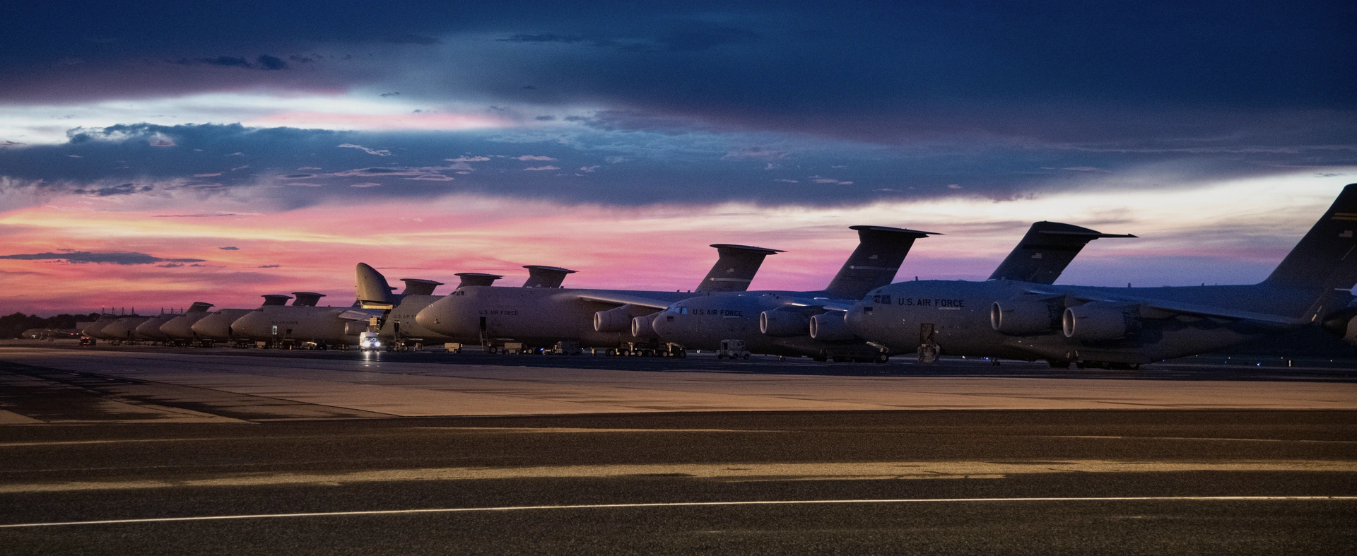A long line of U.S. Air Force C-5M Super Galaxy and C-17 Globemaster III aircraft parked on a flight line at dusk.